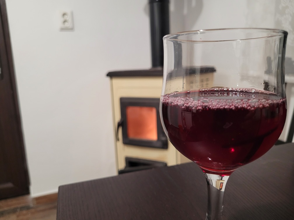 Pahar cu vin de casă roșu, limpede și cristalin, așezat pe masă lângă sobă.